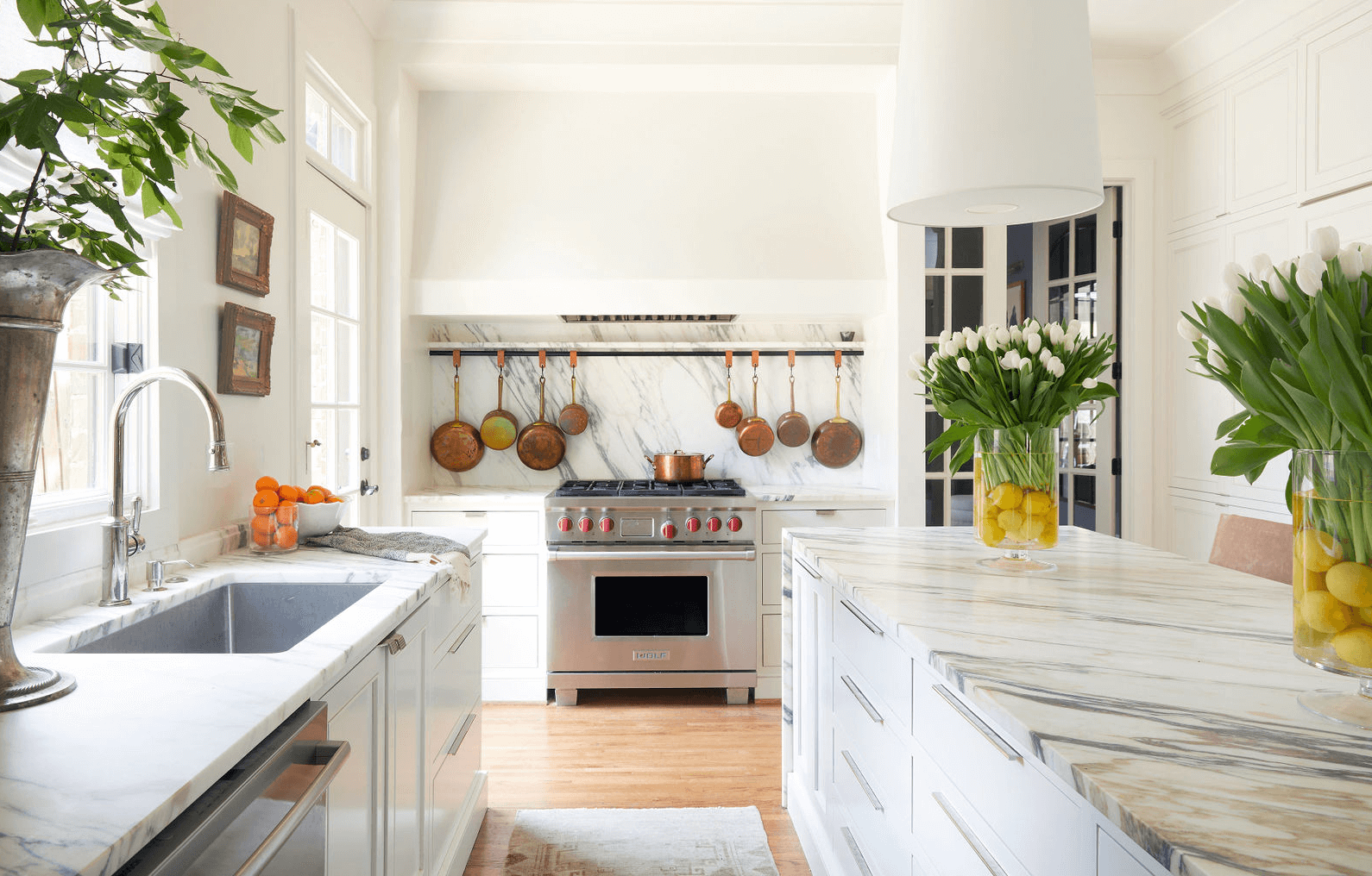 The Top Interior Designers and Architects in Birmingham - 5 A modern kitchen with a marble island, marble countertops, hanging copper pots, a stainless steel stove, and white cabinetry. Designed by some of the best interior designers and architects in Birmingham, the space features vases of white tulips and lemons placed on the island and counter.
